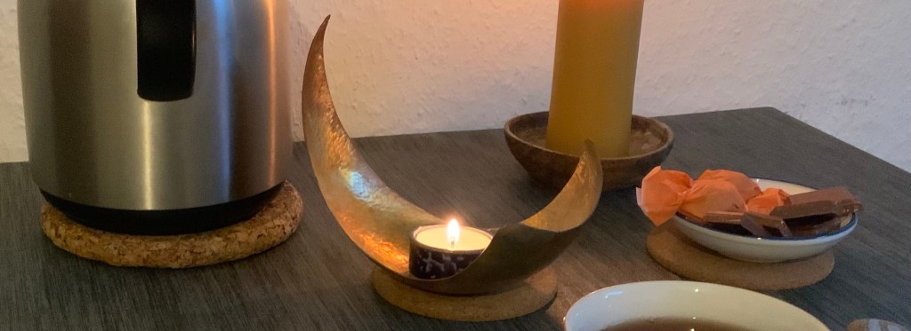 Teekanne und -tasse, ein Schälchen mit Schoki, zwei Kerzen, ein Engel auf einen kleinen Stein gemalt und das Macbook vor mir auf dem Tisch. (Angeschnitten.)