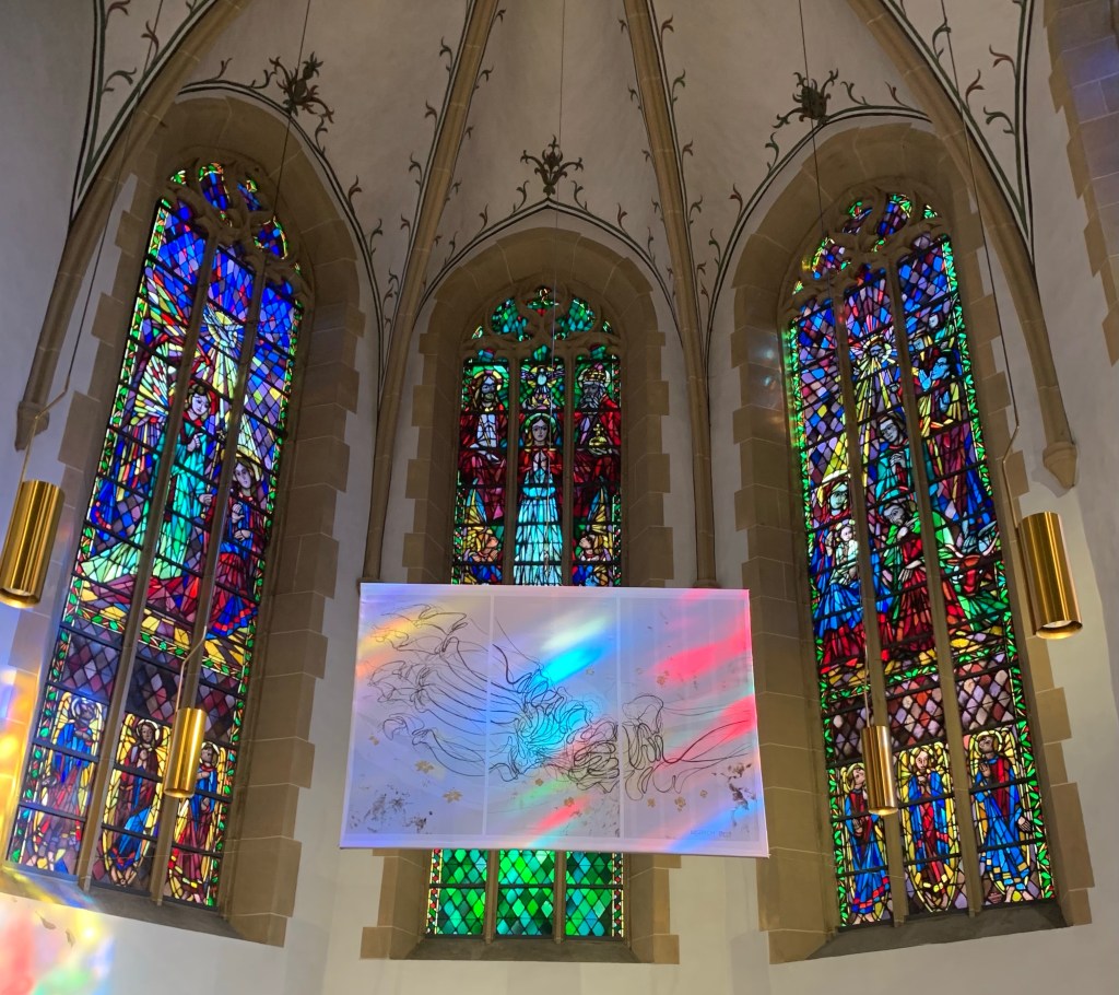 Das Hungertuch, aufgehängt im Altarraum, dahinter drei sehr bunte Fenster.