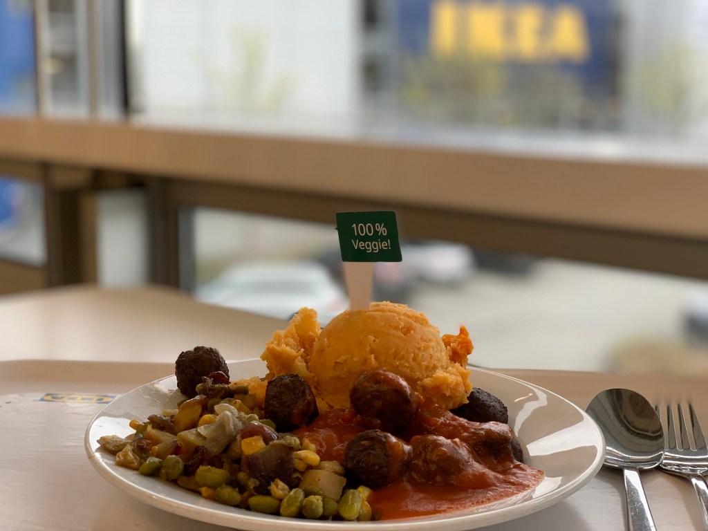 Das Mittagessen bei Ikea auf einem Tablett mit Besteck daneben und einem "100 % Veggie!"-Schildchen in den Püree gesteckt. Im Hintergrund der verschwommene Blick aus dem Fenster, der den gelben Ikea-Schriftzug erkennen lässt.