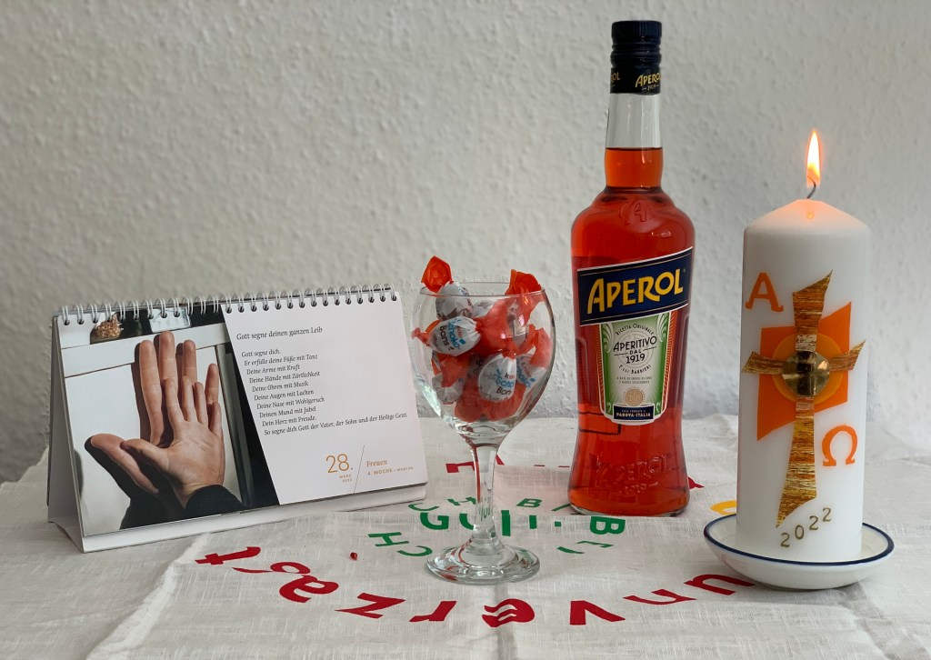 Weiße Leinendecke mit buntem Aufdruck und darauf von links nach rechts: Der Fastenkalender, aufgeschlagen an dem Tag mit dem Segen (Datum in orange); ein Wandglas voll mit Kinder Schokobons (orange), eine Flasche Aperol (orange), die brennende Osterkerze (orange Isignien). 