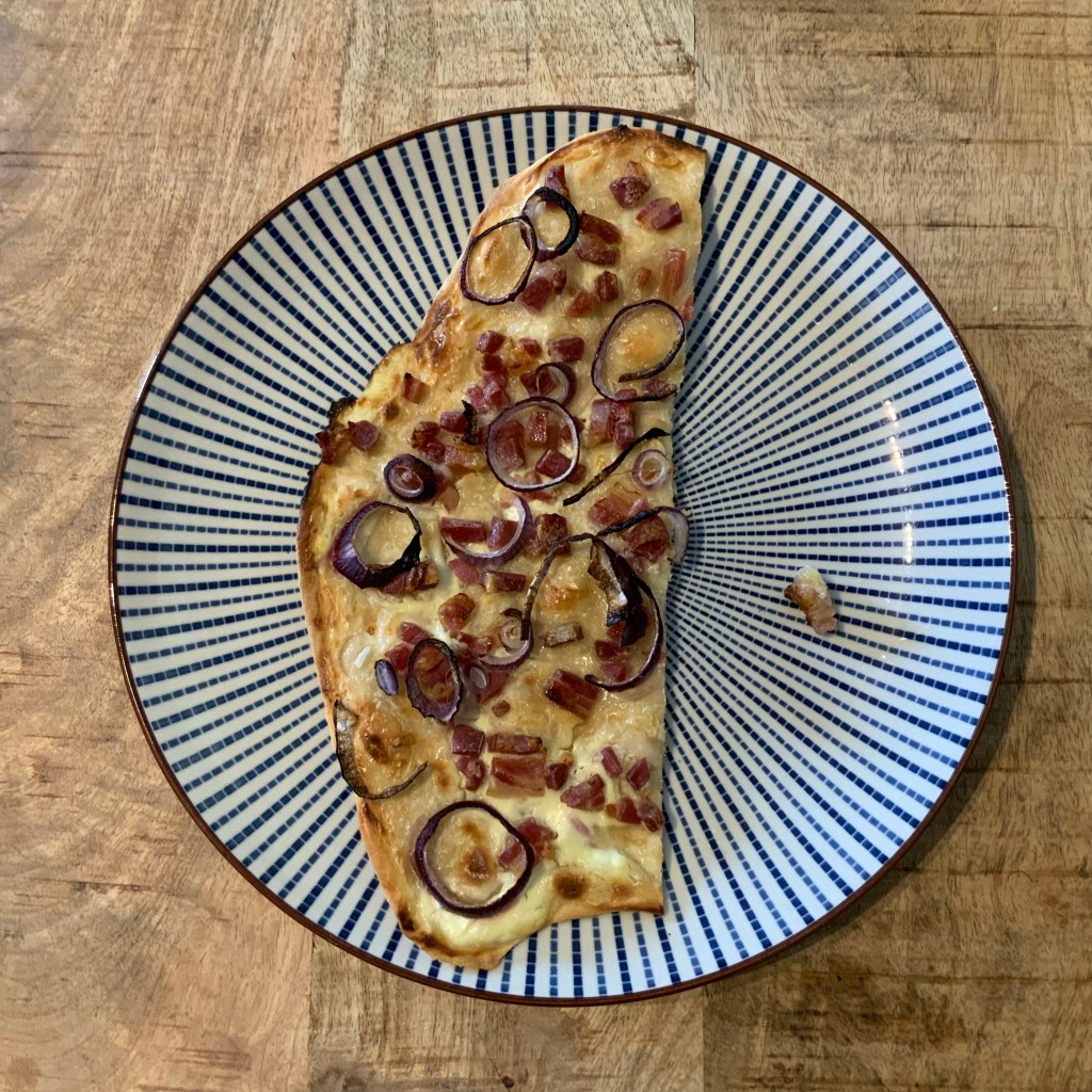 Ein Stück (selbnstgemachter) Flammkuchen auf einem Teller mit weißen und blauen Strahlen bzw. Streifen.