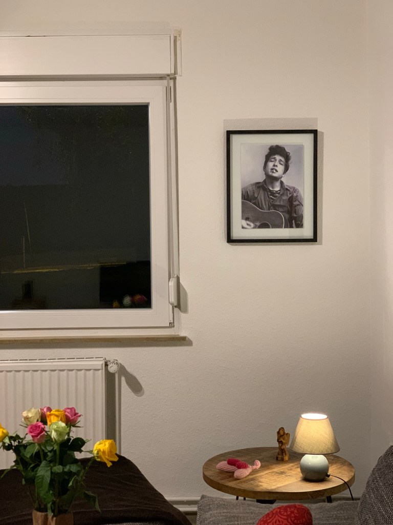 Das gerahmte schwarzweiß Foto vom jungen Bob Dylan, aufgehängt in der Ecke neben dem Wohnzimmerfenster (man sieht, es ist dunkel), davor ein Beistelltischchen, darauf eine Lampe, ein Olivenholzengel und eine gehäkelte Clitoris. Daneben, vermutlich auf einem anderen Tisch, eine Vase mit bunten Rosen und ein Stück der Couch.
