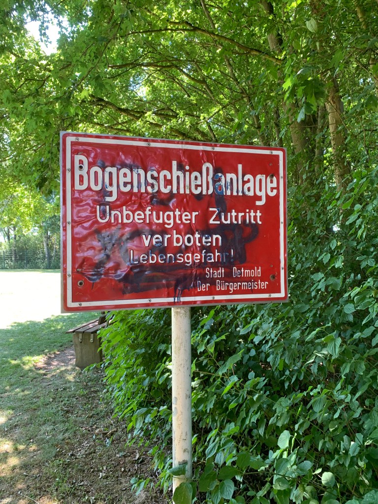 Vor Bäumen und Büschen ein rotes Warnschikd mit weißer Schrift und ein paar Graffitos: "Bogenschießanlage. Unbefugter Zutritt verboten. Lebensgefahr! Stadt Detmild. Der Bürgermeister