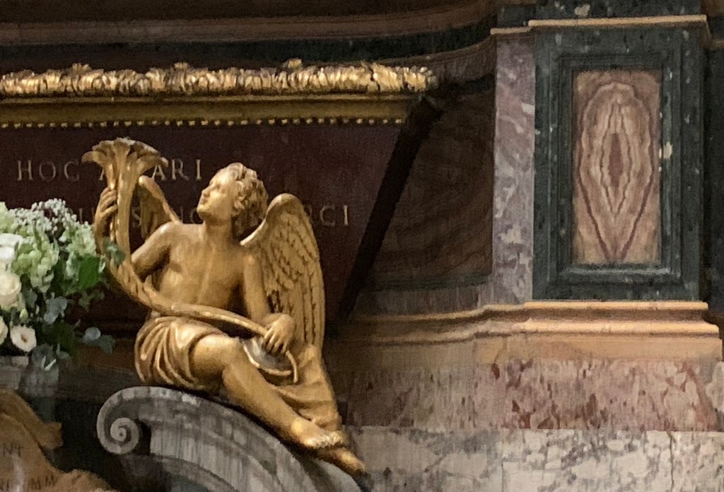 Nahaufnahme der rechten Hälfte des Hauptaltars. Links im Vordergrund Teile eines weißen Blumenbouquets, daneben ein knabenhafter goldener Engel mit einem Ding, das halb Posaune, halb Öiöie ist. Dahinter ein Marmorarkophag mit Reliquien, daneben in die Altarseite eingelassen wie eine Kassette die Umrisse einer geöffneten Vulva, auch aus Marmor.