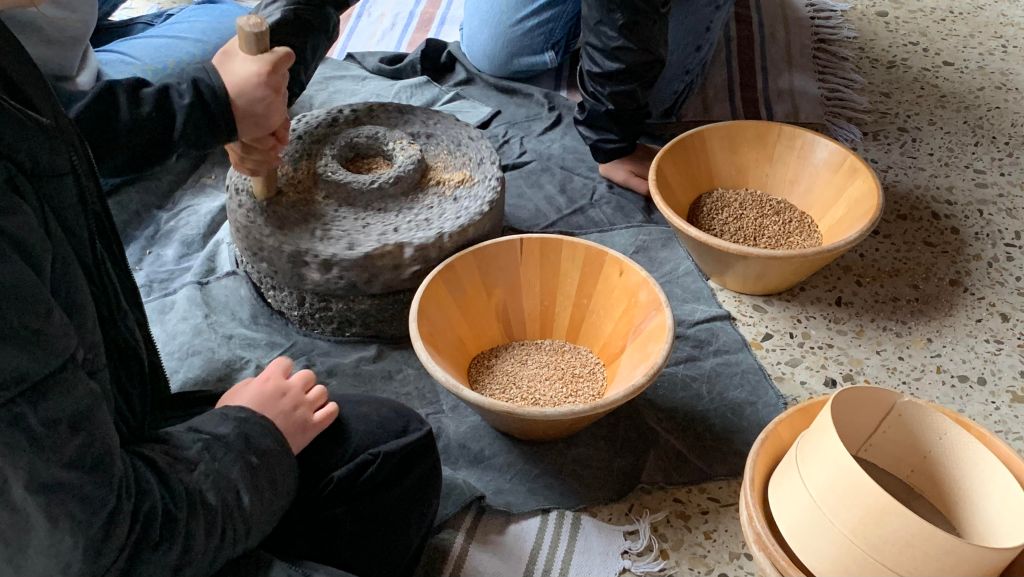 Zwei Konfis (nicht zu erkennen) sitzen auf dem Fußboden und bedienen eine Handmühle (zwei tellergroße Mühlsteine). Daneben Holzschüsseln mit Körnern, Schrot, Mehl und Sieb.