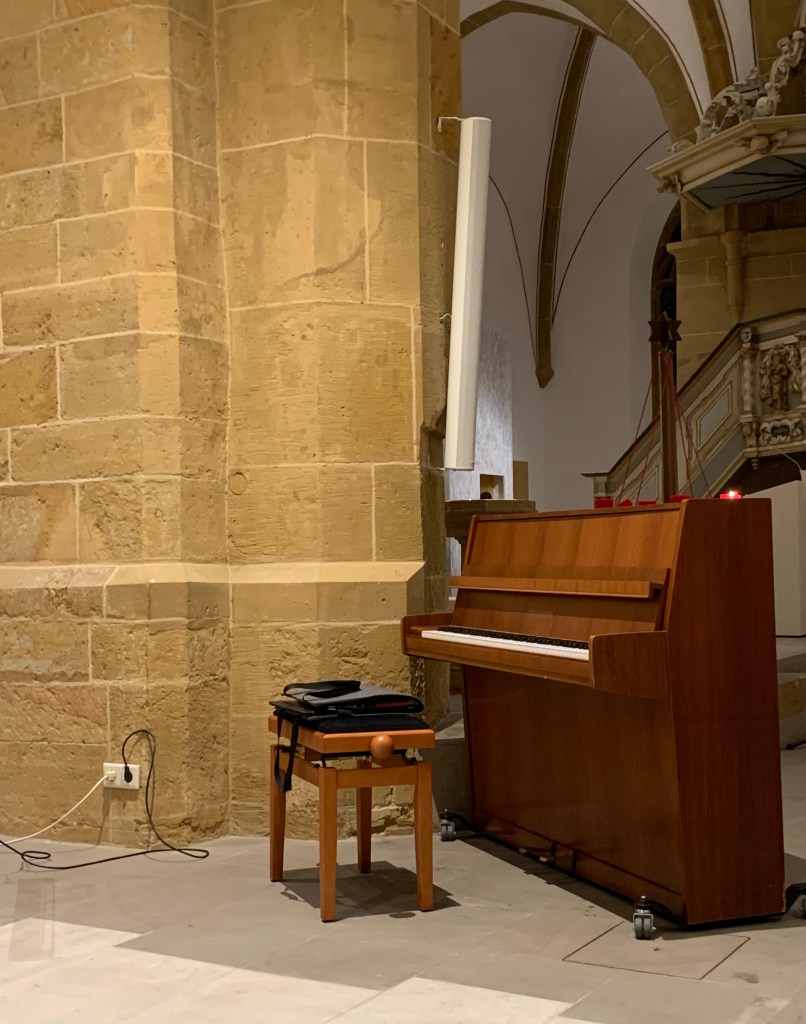 Das Klavier, ein mittelbraunes Grotrian-Steinweg, vor dem Konzert neben einer dicken Kirchensäule mit länglichem Lautsprecher, im Hintergrund die historische Kanzel mit Schnitzwerk. Davor ein Klavierhocker, auf dem der Pianist seine Notentaschen abgelegt hat.