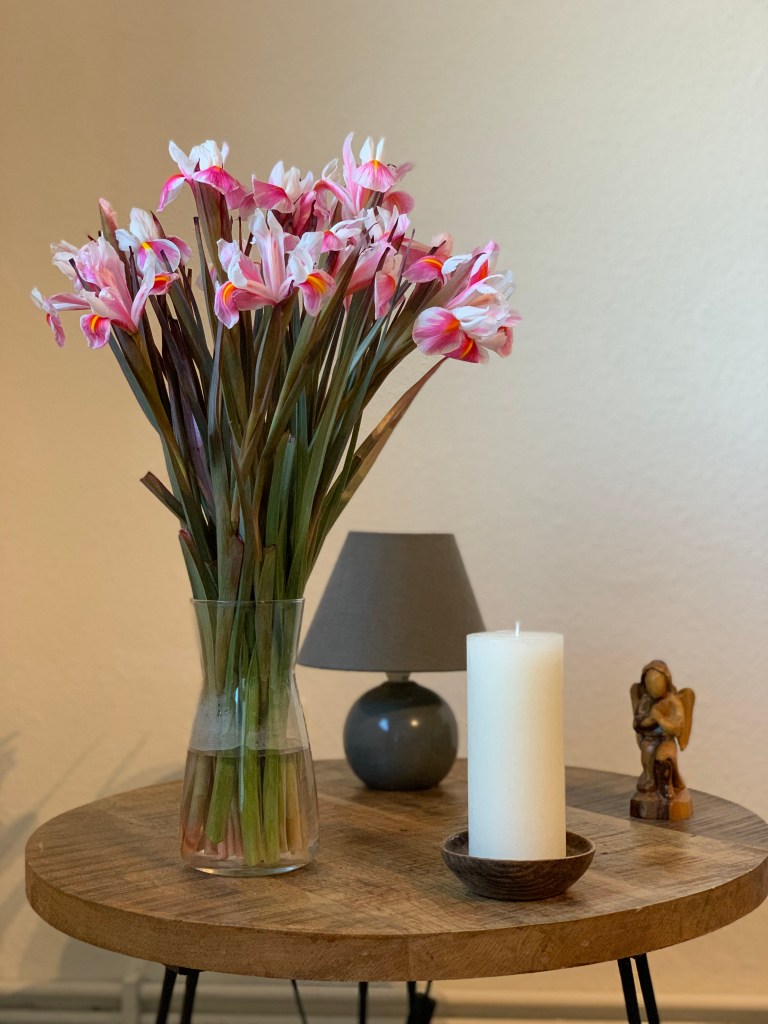 Ein Strauß weißpinker Iris in einer Glasvase auf einem Beistelltisch mit Lämpchen, Kerze (aus) und Olivenholzengel.