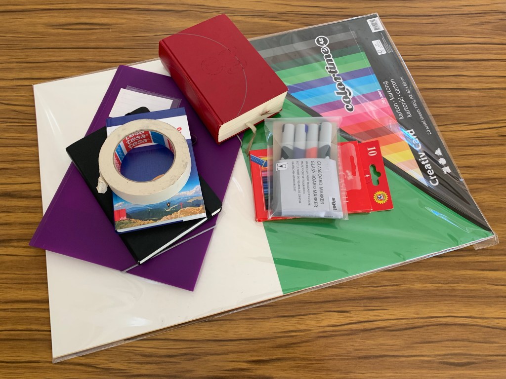 Material für den Katte auf einem Tisch: Fotokarton, Stifte, Kreppband, EG, Lehrbuch, Kladde, Mappe.