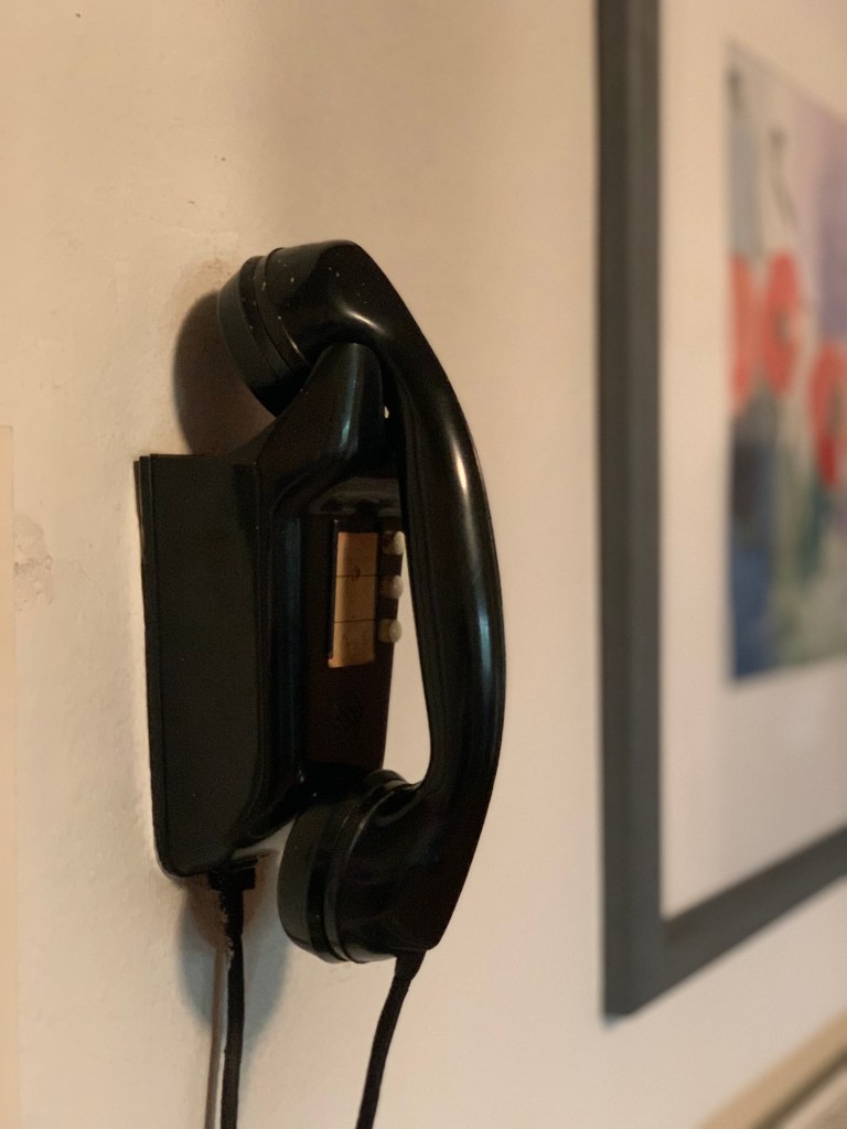 Schwarzes Wandtelefon aus Bakelit. Daneben hängt schemenhaft sichtbar ein Bild mit Blumen.