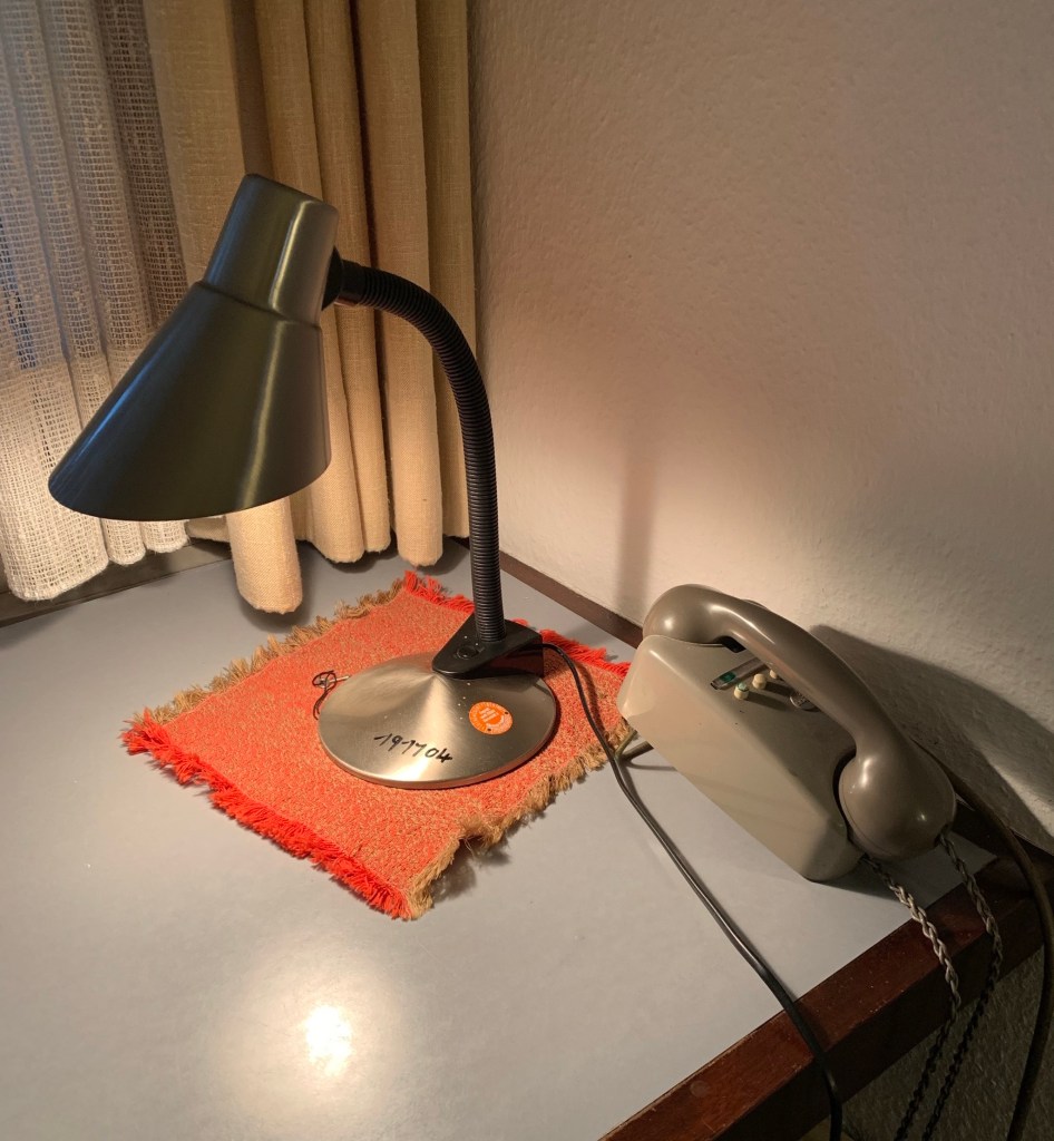 Telefongraues Haustelefon, daneben ein verschossenes rotorangenes Deckchen und darauf eine Tischlampe, alles im Stil der frühen 60er Jahre.