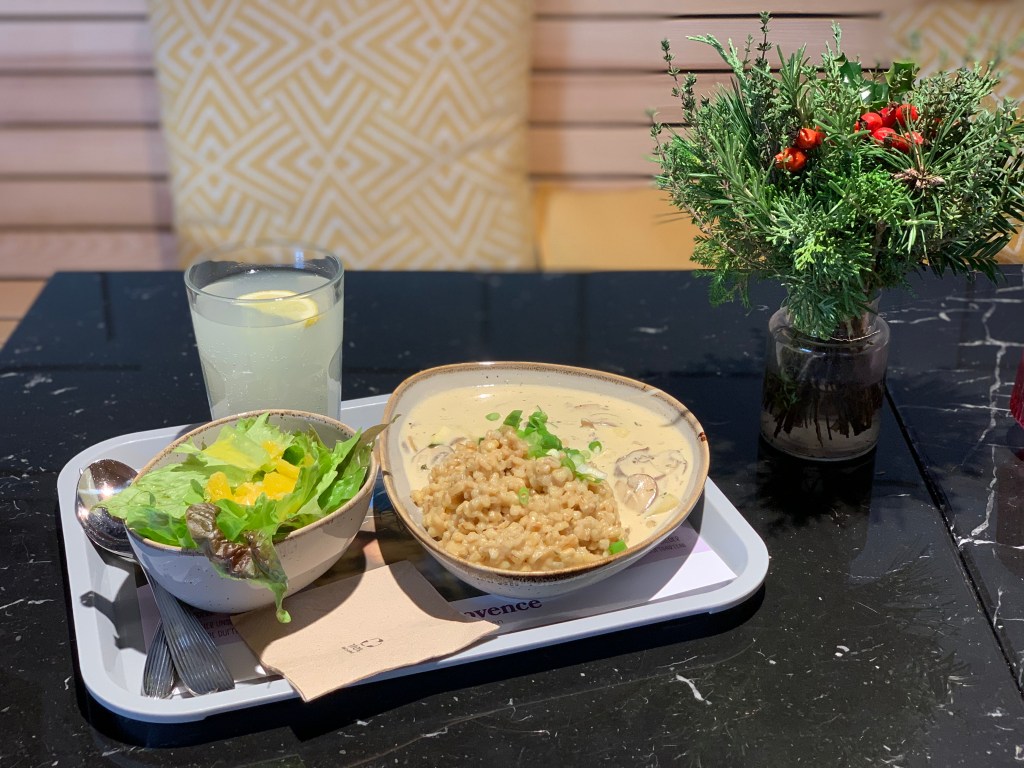 Die Schalen mit dem Mittagessen und das Limoglas stehen mit Besteck auf einem Plastiktablett, das wiederum auf einem Bistrotisch mit shcwarzer Marmortischplatte steht. Daneben ein Dekosträußchen mit Tannengrün und roten Zierbeeren, dahinter eine hölzerne Sitzbank mit Kissen zum Anlehnen.