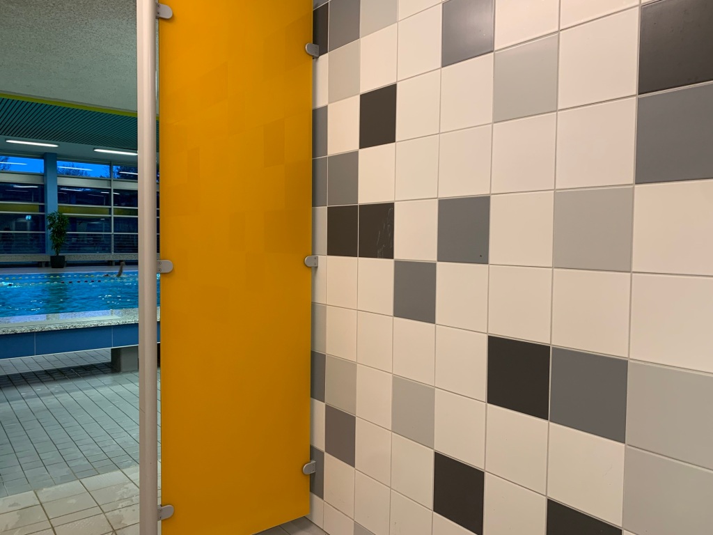 Fliesenwand und Sichtblende im Duschgang geben einen schmalen Blick in die Schwimmhalle auf das Becken frei.