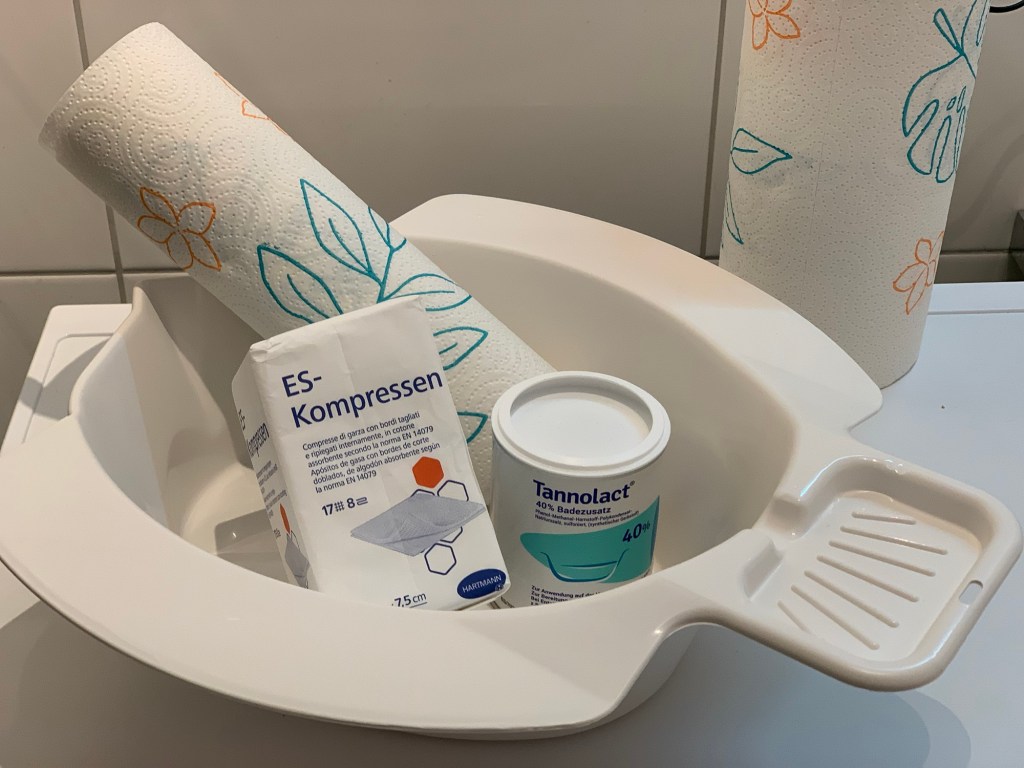 Weiße Schüssel für Sitzbäder in Form eines Fisches, darin Küchenrolle, Kompressen und Tannolact medizinischer Badezusatz.
