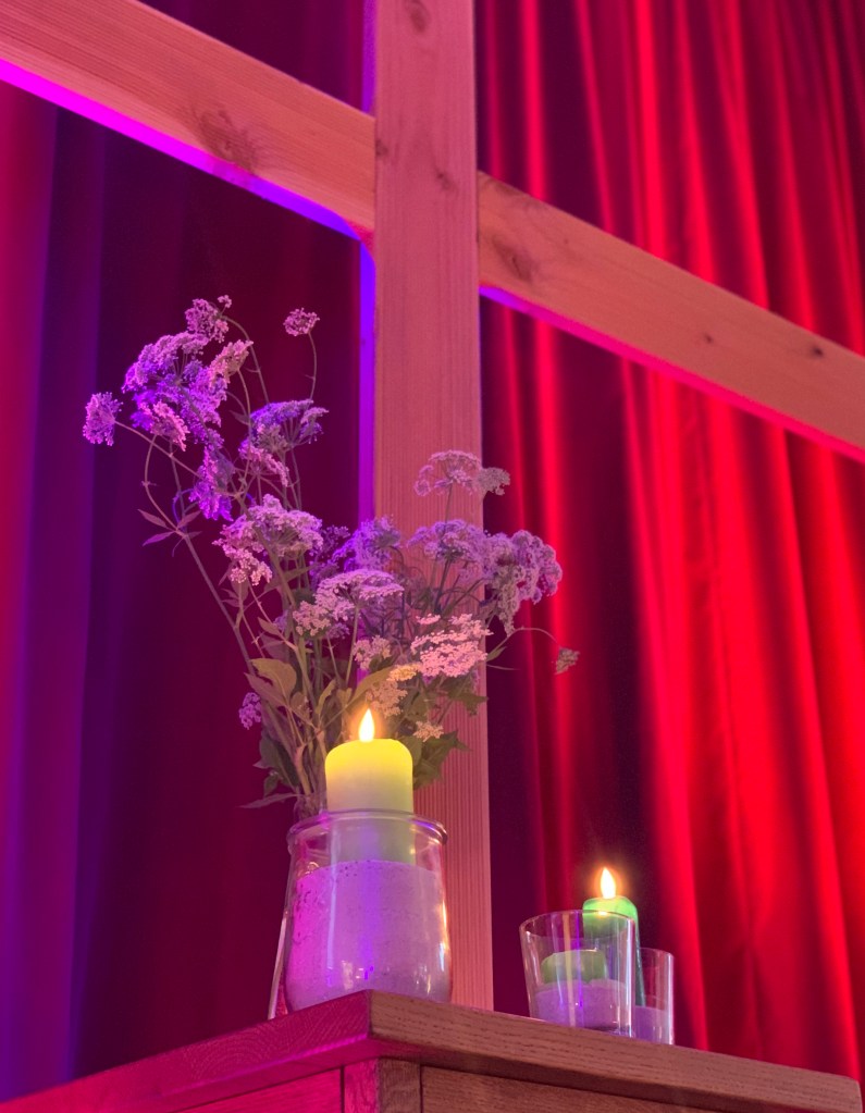 Holzkreuz mit einem Holztisch davor, auf dem Blumen und brennende Kerzen stehen. Das Ganze wird mit rotem und violettem Licht angestrahlt, sodass eine schöne, kitschig-moderne Atmosphäre entsteht.