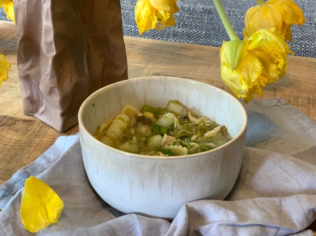 Eine Schale Nudesuppe dekorativ unter fast verblühten gelben Tulpen und auf einer hellgrauen Stoffserviette angerichtet.