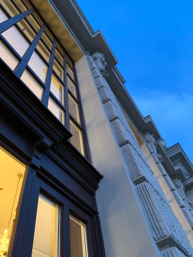 Vom Balkon die Fassade hoch fotografiert. Warmes Licht scheint durch große Kassettenfenster. Man sieht, dass die Fassade irgendwie historisch ist, aber viel mehr erkennt man nicht. (Es ist eine Mischung aus Jugendstil und Neobarock.)