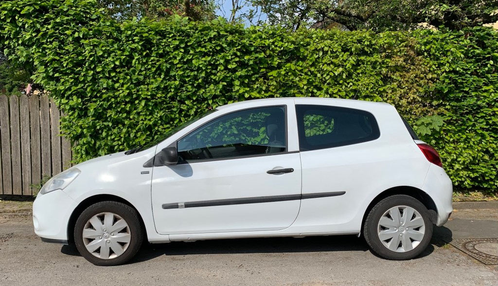 Der DeLorean (ein gut zehn Jahre alter weißer Renault Clio) von der Seite vor einer Laubhecke im Sonnenlicht, sodass man die Dellen in der Tür kaum sieht.