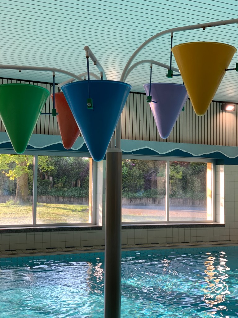 Foto des Lernschwimmbeckens. Im Vordergrund an einem Ständer, dick wie ein kleiner Laternenpfahl und auch recht hoch, eine Art Mobile aus großen, bunten Trichtern. (Es ist jetzt ausgeschaltet, in Betrieb kippen sich die Trichter reihum Wasser weiter und irgendwann aus.)