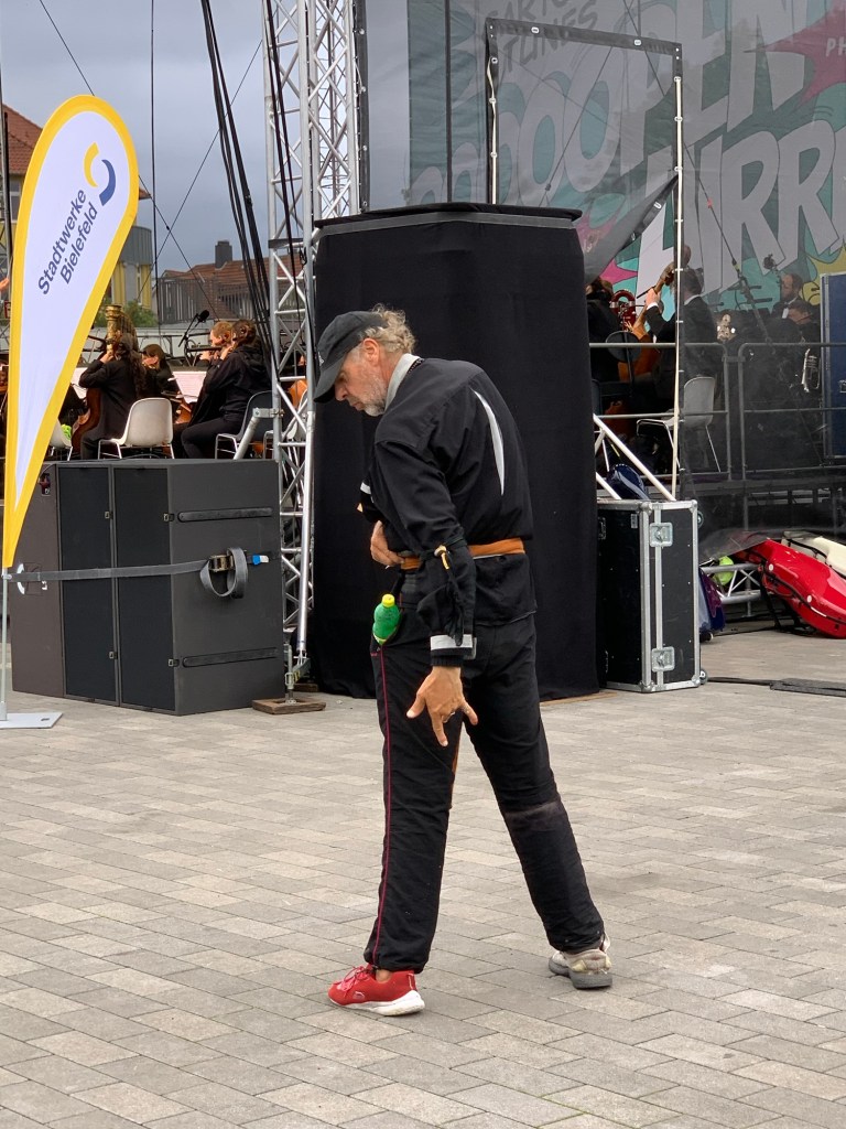 Der tanzende Mann vor der Bühne, das Orchester im Hintergrund. Er ist groß, schwarz gekleidet, mit Cappy und einem roten und einem weißen Turnschuh.