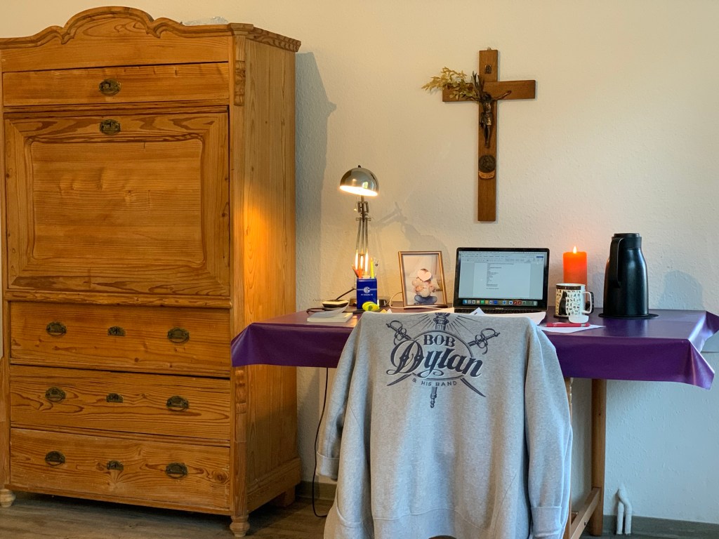 Neben meinem Sekretär (antik, Kiefer) mein privater Schreibtisch: Mit Kruzifix mit Corpus und Palmzweig, darüber, Macbook, Teetasse und -kanne und brennender Kerze. Außerdem eine Stiftebox von Schalke 04 und ein Foto der Enkelin, auf deren Lächeln ich fürs Foto ein großes rosa Herz geklebt habe, damit man sie nicht erkennt. Vor dem Tisch ein Stuhl, über dem eine graue Sweatshirtjacke mit Bob-Dylan-Rückenaufdruck hängt.
