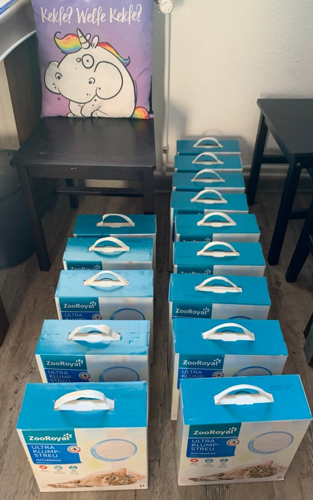 14 Kartons Katzenstreu zu sechs Litern, überwiegend in Zweierreihen. Im Hintergrund ein Stuhl, ein Tischchen und ein Stück Papierkorb. Auf dem Stuhl steht ein fliederfarbenes Zierkissen mit einem Regenbogeneinhor. drauf, das mit vollen Backen kaut. Darüber die Worte: "Kekfe? Welfe Kekfe!"