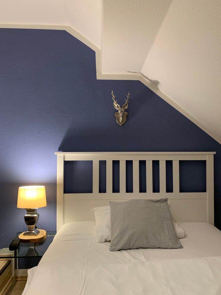 Hotelbett in weißem Landhausstil. Dahinter eine blaue Wand mit einem kleinen Hirschkopf aus Metall. Daneben die Nachtischlampe.