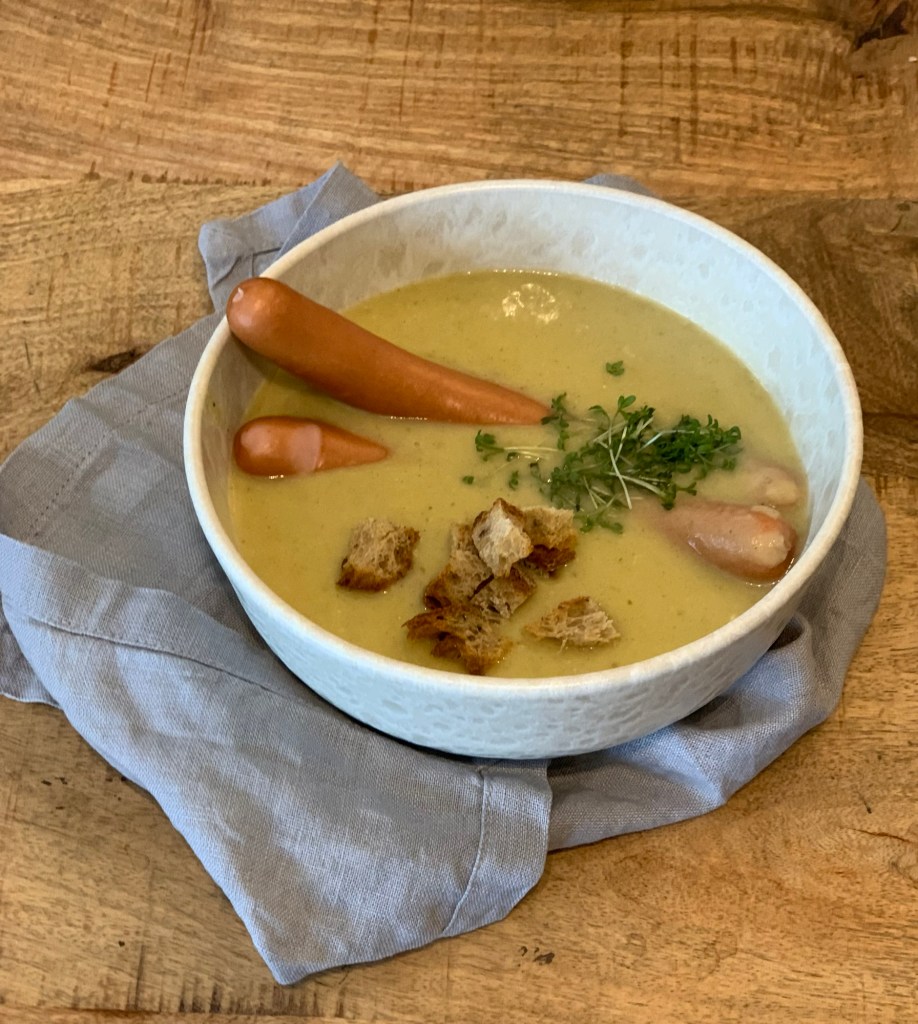 Eine Schale Kartoffelsuppe mit zwei Bockwürstchen darin, etwas Kresse und ein paar Croutons.