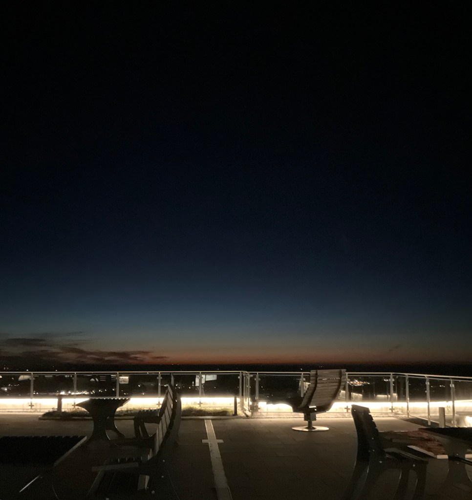 Blick im Dunkeln auf die Dachterrasse: Sie ein bisschen wie eine kleine Landebahn mit Stühlen aus und ist umrandet mit einem verglasten Geländer. Das ist illuminiert, dazu sieht man die Lichter der Siedlungen dahinter und der Spiegelungen in den Glasscheiben. Über all dem wölbt sich der große, schwarze Himmel mit einem Rest Abenddämmerung am Horizont.