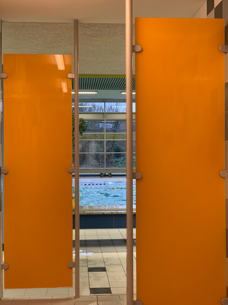 Im Schwimmbad aus dem Umkleidegang Richtung Becken fotografiert. Wobei man vom Becken in der Mitte nur ein wenig sieht, weil rechts und links zwei orangene Trennwände den Blick rein und raus versperren.