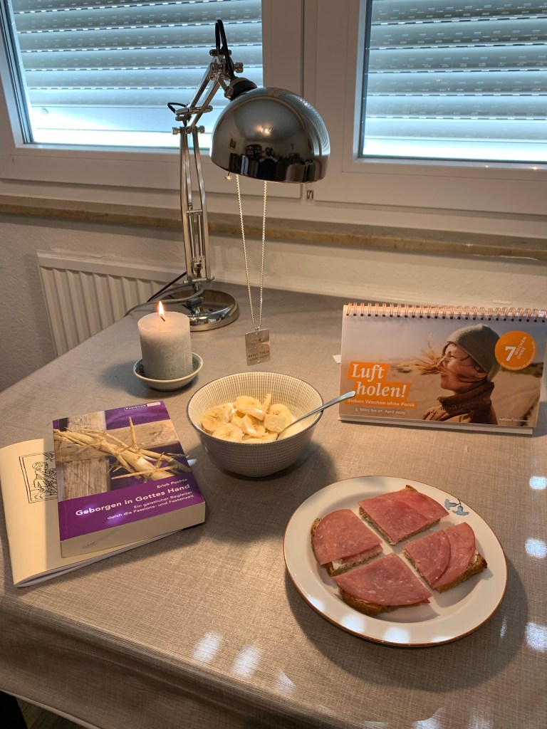 Das Geraffel zu Beginn der Fastenzeit auf meinem Schreibtisch mit der Schreibtischlampe im Hintergrund an der meine Israel-Plakette hängt (Bring thgem home now.). Auf dem Schreibtisch liegen bzw. stehen: Fastenbücher und -kalender, ein Teller mit dem Brot, eine Schale mit Banane (kleingeschnitten) und Joghurt, eine brennende Kerze.