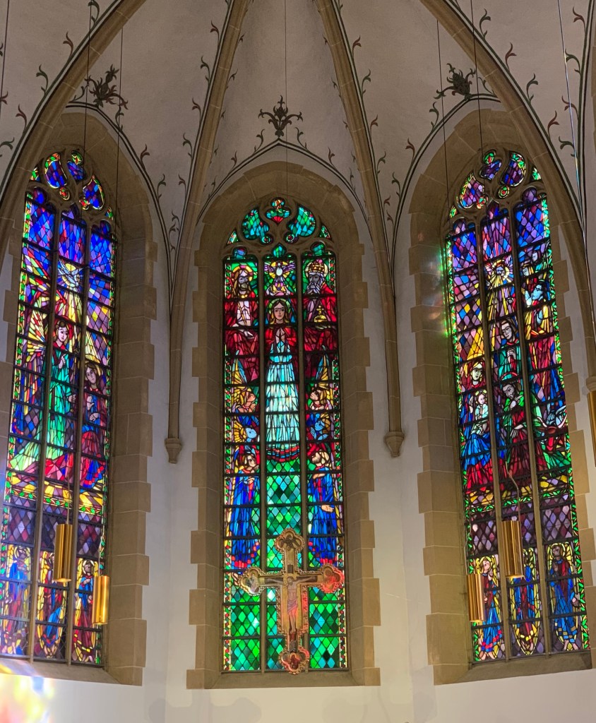 Drei sehr bunte gotische Fenster in der Apsis. Davor hängt ein goldenes Kruzifix.