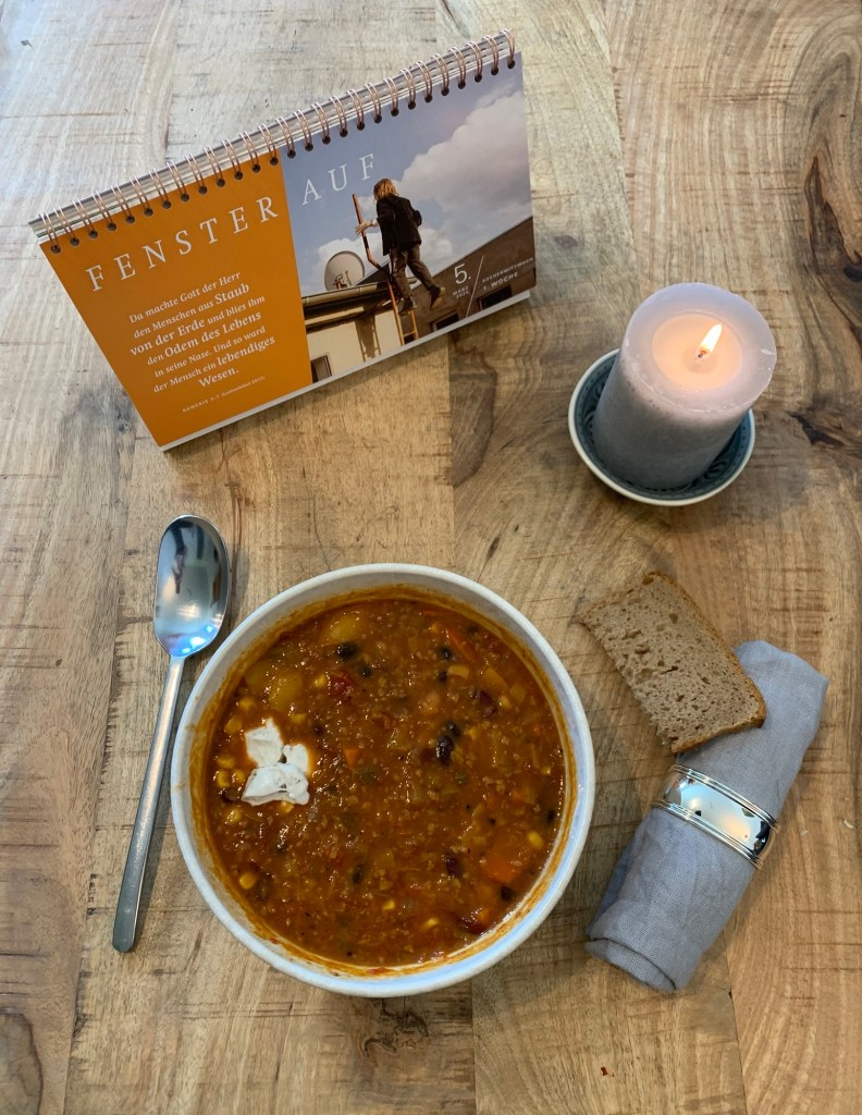 Eine Schale Chili wie beschrieben, drumherum der Kalender von 7 WO, eine Stoffserviette, das Brot, ein Löffel und eine brennende Kerze.