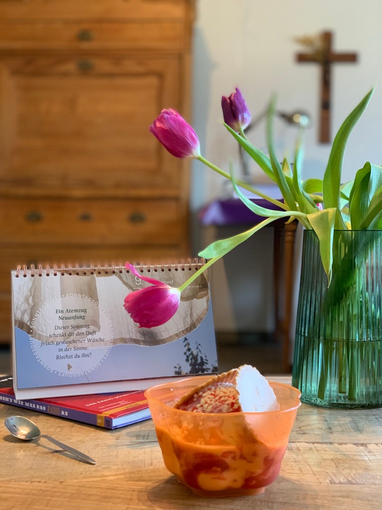 Das Spaghettieis mit den runden Waffeln zum Abdecken oben drauf auf meinem Wohnzimmertisch, mit einem Teelöffel, einem Buch, dem Fastenkalender und einer Vase mit Tulpen drumherum. Im Hintergrund mein Sekretär und das Kruzifix an der Wand.