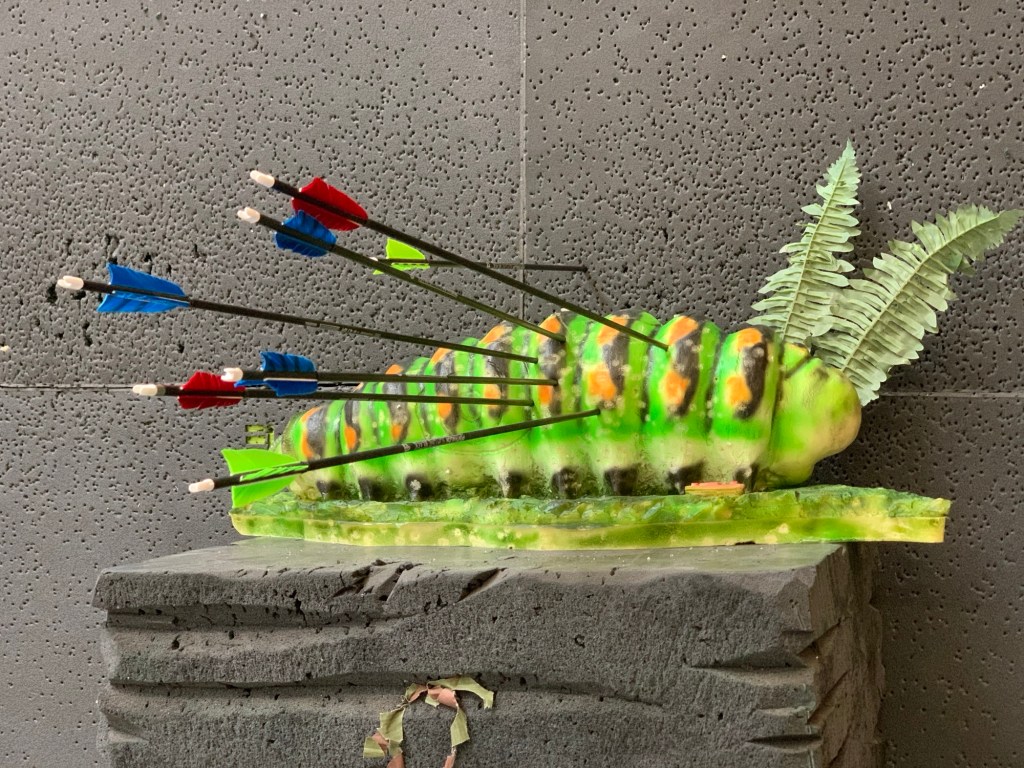 Eine grüne Gummiattrappe einer Schmetterlingsraupe als Schussziel, groß wie ein Oberschenkel. Hinter ihrem Kopf stecken zwei Plastikfarnblätter, die fast wie Fühler aussehen. Die Raupe sitzt auf einem grünen Untergrund und damit auf einem schwarzgrauen Schaumstoffpodest vor einer ebensolchen Wand. In ihr stecken sechs Pfeile, der siebte steckt in der Wand.