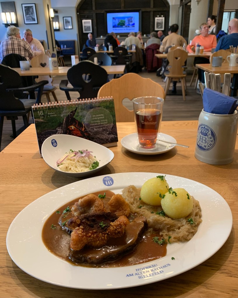 Vor mir auf dem Tisch ein ovaler Teller mit Emblem: Hofbräu am Alten Rathaus Bielefeld. Darauf zwei Scheiben Schweinebraten, drei Stück geröstete, krosse Schwarte, zwei Knödel auf Sauerkraut. Dahinter auf dem Tisch der Fastenkalender, am Sonntag Reminiscere aufgeschlagen und fürs Foto mitgeschleppt; ein Schälchen Krautsalat aus gehobeltem Kraut; ein Glas Assamtee; ein Bierhumpen mit Servietten und Besteck. Im Hintergrund der Blick ins Lokal: Leute an Tischen.
