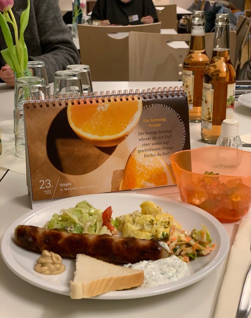 Auf dem gedeckten Tisch im Gemeindehaus vor mir mein Teller mit Bratwurst, Senf, Tzatziki, kleinen Salatportionen und einer Ecke Toast. Dahinter der wieder fürs Foto mitgeschleppte Fastenkalender. Daneben die Gummibärchen, die ich in den orangenen Plastikbecher vom Spaghettieis an Invokavit umgefüllt habe, um sie anbieten zu können.
