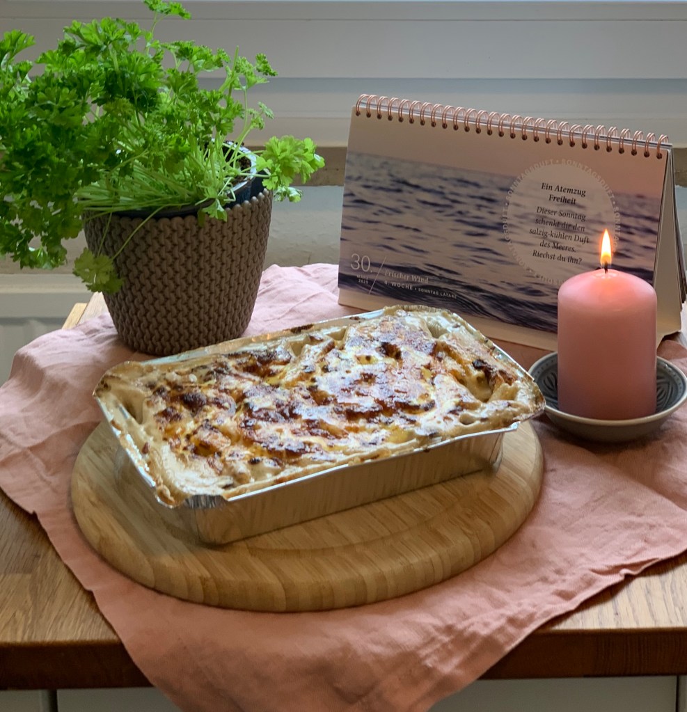 Der Auflauf in der Aluschale auf einem Holzbrett. Darunter eine altrosa Stoffserviette, daneben eine brennende rosa Stumpemkerze. Links dahinter eine Topfpetersilie und rechts der Fastenkalender, ein Motiv mit Meereswellen und zartem Rosa (Morgenrot).