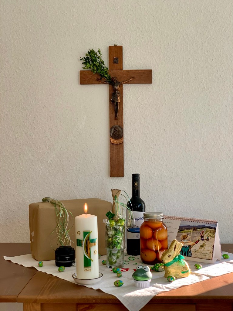 Blick auf meinen privaten Schreibtisch, gedeckt als österlicher Gabentisch, darüber das Kruzifix mit dem neuen Palmzweig. Als Ostergaben: Ein kleines Paket in Packpapier mit grün-silber-goldenem Bast-Schleifen-Büschel; ein Canon-Objektiv 50 mm; die Osterkerze mit grün-goldenem-Kreuz; ein Glas mit Süßkram und einem stiftartigem Gechenk, ebenfalls wie geschildert verpackt; kleine Schokieier, der ganze Süßkram in grün-gold, ein Lindhase (sitzend) in gold mit grüner Schleife; der Fastenkalender auf dem Tag mit dem Gedicht von Virginia Satir; eine Flasche Dornfelder trocken; ein Glas selbstgemachte Soleier; eine kleine Nagelbrüste mir einem grünen, niedlichen Krokodilkopf drauf. Eine weiße Leinentischdecke mit Aufdruck. 