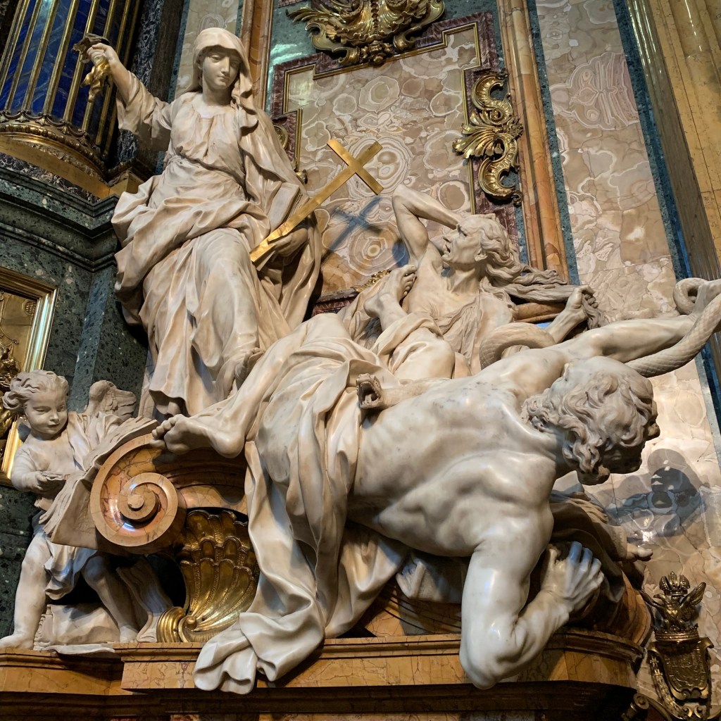 Maria (?) mit einem Kreuz bewaffnet und eine Putte kämpfen gegen die Häresie, die aussieht wie Melanachthon und ein weiterer Mann, als wären sie vom Olymp gestürzt. Unter dem Mann lugt ein Buch hervor. Man sieht es hier nicht, aber es steht sinngenmäß NT von Luther drauf. Alles aus Marmor, Kreuz in gold.