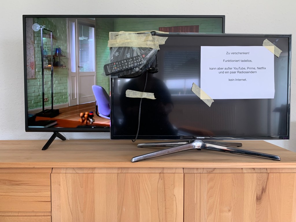 Der eingeschaltete neue Fernseher auf der dafür vorgesehenen Kommode. Davor steht der alte Fernseher, etwas kleiner und fertig zum Abtransport: Die Fernbedienungen in einem Gefrierbeutel mit Krepp oben an der Ecke festgeklebt, ebenso das Stromkabel. Außerdem habe ich ein Blatt ausgedruckt und drangeklebt mit folgendem Text: "Zu verschenken! Funktioniert tadellos, kann aber außer YouTube, Prime, Netflix und ein paar Radiosendern kein Internet."