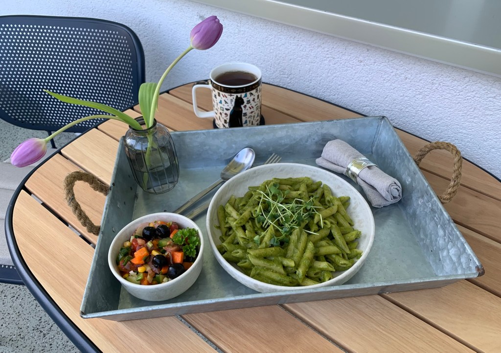 Auf meinem Balkontisch steht meine Katzentasse mit Tee und davor ein Metalltablett. Darauf eine kleine Rauchglasvase mit zwei helllila Tulpen, Besteck und eine Stoffserviette im Serviettenring, ein Teller Nudeln mit Pesto und ein paar Ziersprossen und ein Schälchen Salat.