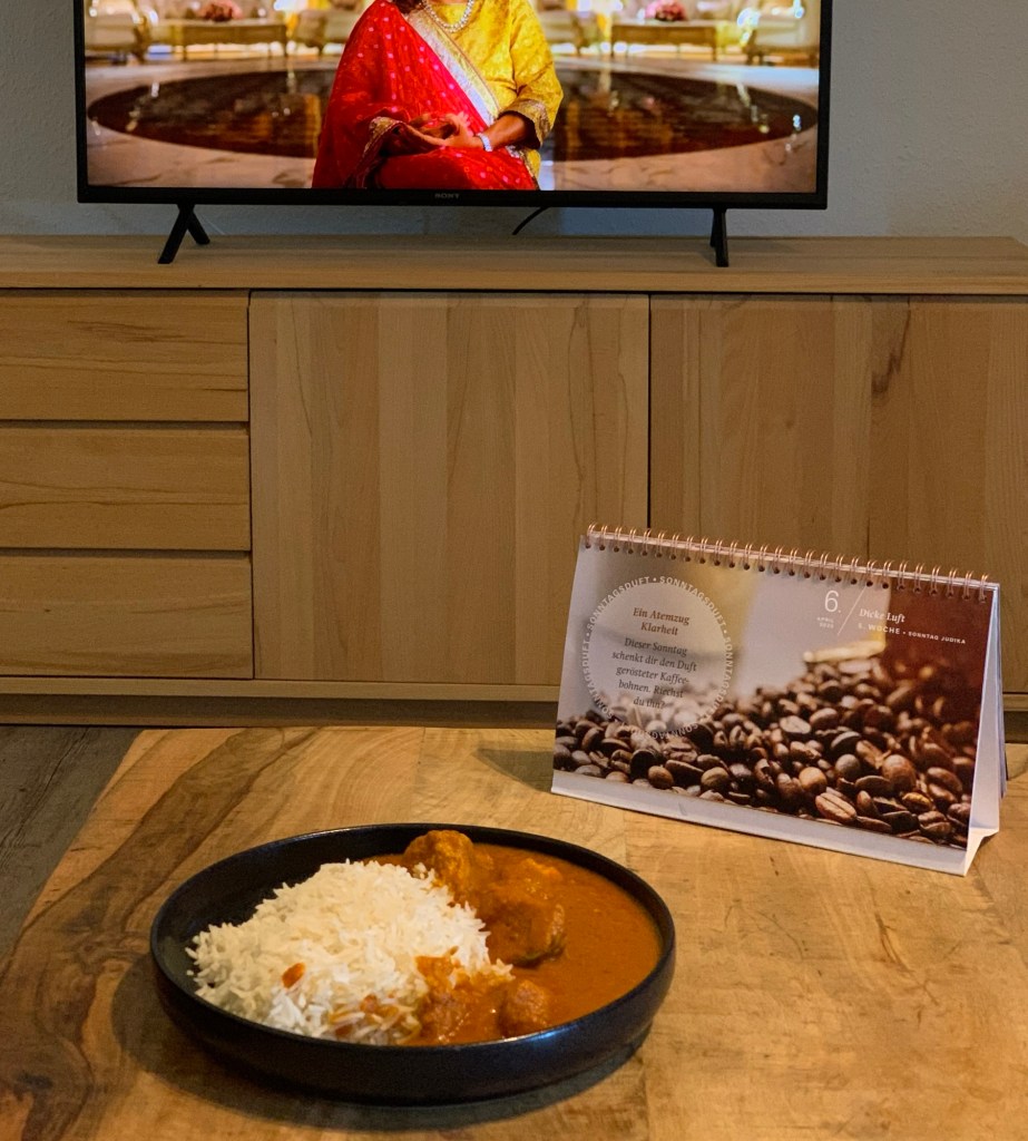 Auf dem Wohnzimmertisch ein schwarzer Teller mit Butter Chicken und Reis, daneben der Fastenkalender mit einem Motiv über den Duft von Kaffeebohnen. Es sind auch viele Kaffeebohnen abgebildet. Im Hintergrund der Fernseher mit Sima Taparia von Indien, Matchmaking im Bild. Ich habe ihr allerdings den Kopf abgeschnitten, damit man mir wegen der Bildrechte keinen Strick dreht.