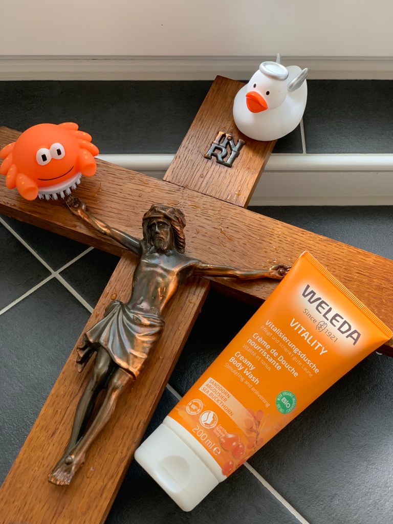 Ein Kruzifix aus braunem Holz mit Kupfercorpus und benetzt mit einigen Wassertropfen. Auf dem linken Querbalken sitzt ein orangener Krebs, der eine kleine Nagelbürste ist. Über Jesu Häupten sitzt eine kleine, weiße Quietscheente mit orangenem Schnabel, silbernem Heiligenschein und auf dem Rücken zwei zusätzlichen silbernen Engelsflügeln. An dem rechten Balken lehnt eine Tube orangenes Duschgel. 