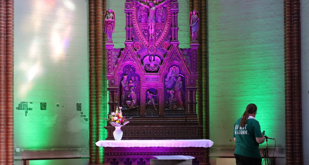 Blick auf den Altar in der Apsis, der grün-violett angeleuchtet ist. Auf dem Altar steht ein Blumenstrauße, daneben jemand vom Technikteam mit dem Rücken zum Bild.