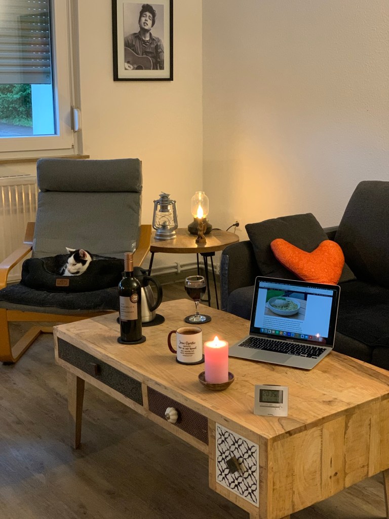 Blick auf die Sitzecke in meinem Wohnzimmer. Auf dem Pöng (Sessel) der #DiätKater in seinem Liegebett. Dahinter ein Stück vom Fenster, daneben das gerahmte Foto des jungen Bob Dylan. Darunter ein Beistelltsich mit der Petroleumlampe (aus) und einer Vintage-Stromleuchte (an). Daneben das Sofa mit einem Herzkissen. Davor der Wohnzimmertisch. Darauf stehen: Eine Teekanne- und Tasse; eine Weinflasche plus Glas; eine rosa Stumpenkerze (an); das Macbook mit diesem Post; ein Wecker statt Wohnzimmeruhr.