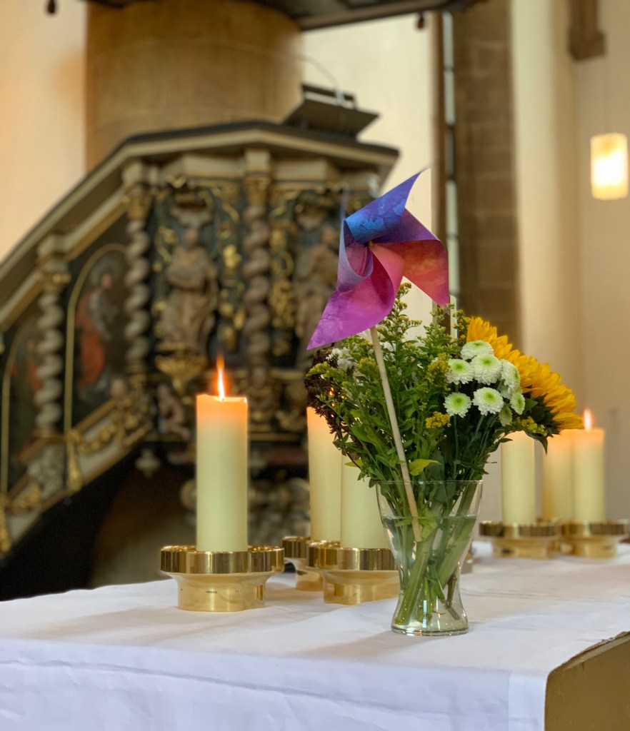 Der Altar beim CSD-Gottesdienst mit Kerzen und Blumenstrauß, in dem ein kleines Windrad in rot, lila, rosa, blau steckt. Im Hintergrund die Kanzel, am oberen Bildrand sieht man ein wenig, dass sie einen Schalldeckel hat.