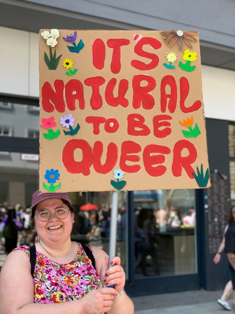 Eine lächelnde, mollige Frau in rot-geblümtem Kleid hält ein Schild hoch. Darauf sind ebenfalls Blumen und es steht in großer roter Schrift: "It's natural to bei Queer."