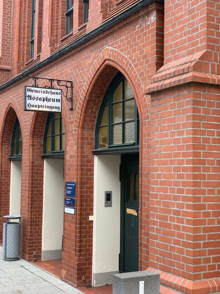 Eingang eines Backsteingebäudes mit neogotisch spitz zulaufenden Türen. Darüber ein Emailleschild in Fraktur: "Gemeindehaus Assapheum Haupteingang".