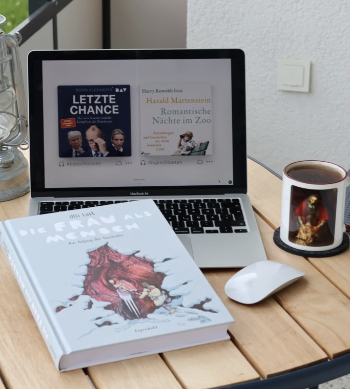 Vor mir auf dem Balkontisch: Das Macbook mit den Titelbildern von Alexander und Martenstein. Davor, halb auf der Tastatur, das Printbuch von Lust. Daneben die Maus und eine Tasse Tee mit dem verlorenen Sohn von Rembrandt drauf. Hinten links sieht man ein Stückchen Petroleumlampe.