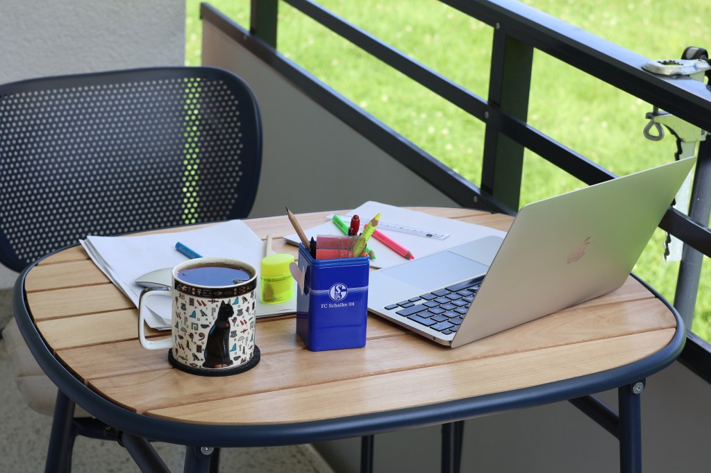 Foto auf meinen Balkontisch, aber nicht vom Stuhl, sondern von der anderen Seite aus. Man sieht beim aufgeklappten Macbook also von der Rückseite Richtung Schreibsachen statt umgekehrt. Daneben mein privater Stiftecontainer von Schalke 04 und meine randvolle Katzenteetasse. Davor liegen DIN A4 Blätter und darauf Textmarker-Buntstifte, Maus, Bleistift, Lineal, Anspitzer. Dann sieht man meine Stuhllehne, einen Teil des Balkongeländers und den Rasen davor.